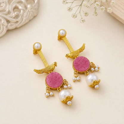 : Pair of Rosy Sparrow anti tarnish brass earrings with pink round stones, gold-toned bird motifs, and pearl danglers on a beige background.