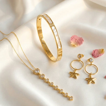 Gold jewelry set with a bangle, heart-accent necklace, and star charm earrings on a soft light fabric background.