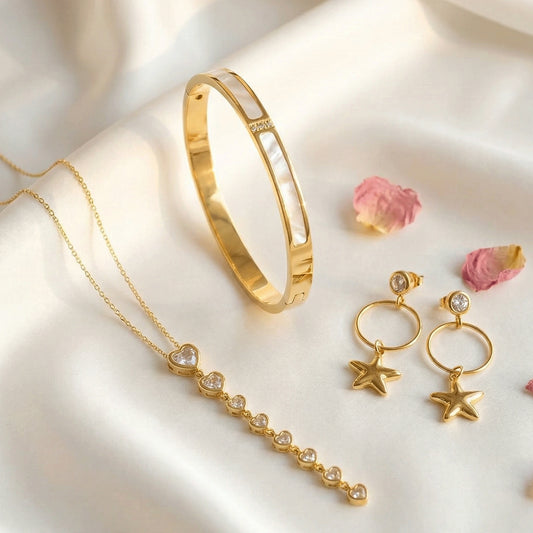 Gold jewelry set with a bangle, heart-accent necklace, and star charm earrings on a soft light fabric background.