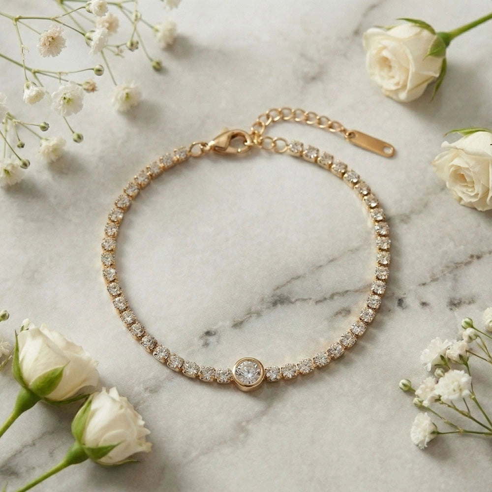 Gold stone tennis bracelet with a central round solitaire charm displayed on a marble surface with white flowers.