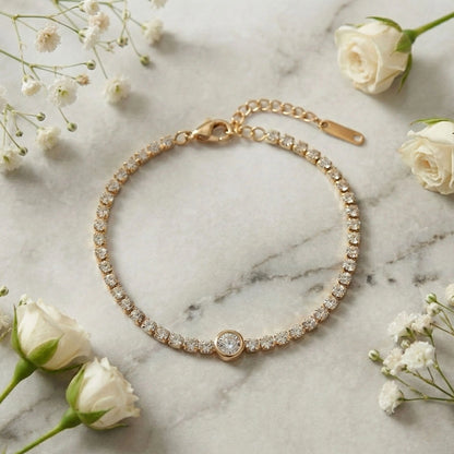 Gold stone tennis bracelet with a central round solitaire charm displayed on a marble surface with white flowers.