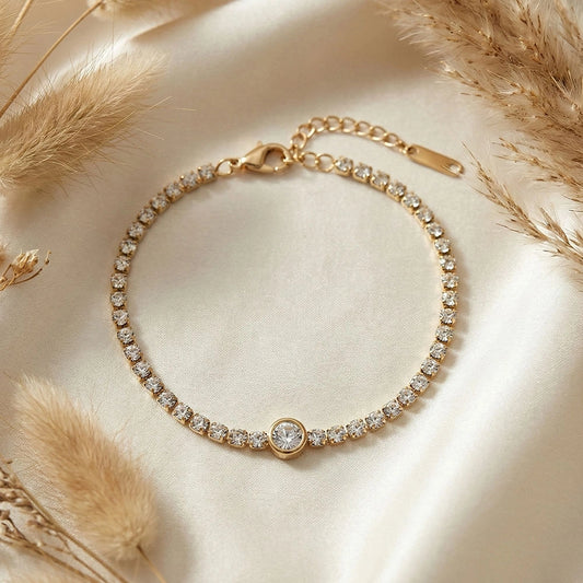 Gold anti-tarnish stone tennis bracelet with central solitaire detail laid on soft champagne fabric with dried florals.