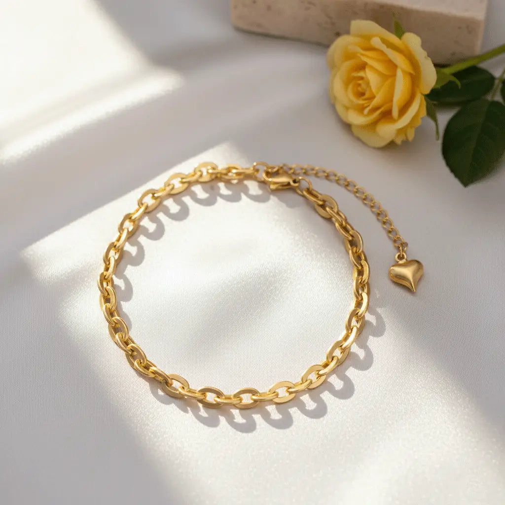 A gold chain bracelet with an adjustable extender and a small heart charm, displayed on soft white fabric with a yellow rose placed beside it.