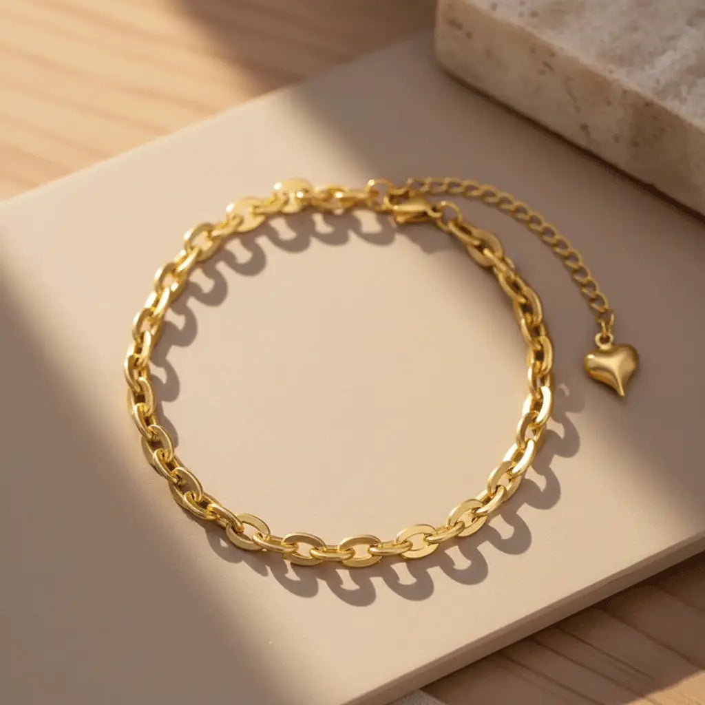 A gold chain bracelet with an adjustable extender and a small heart charm, displayed on a tile.