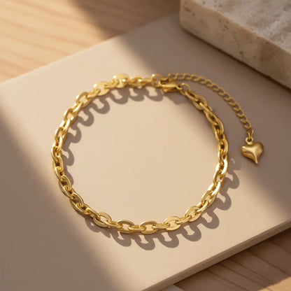 A gold chain bracelet with an adjustable extender and a small heart charm, displayed on a tile.