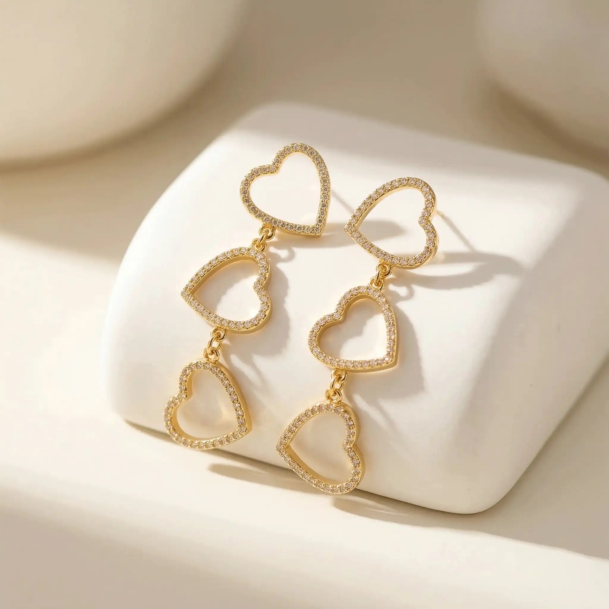 Pair of Three-Tier Heart Earrings with pavé-style stones, arranged on a smooth white geometric block with soft shadows.