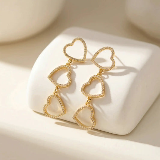 Pair of Three-Tier Heart Earrings with pavé-style stones, arranged on a smooth white geometric block with soft shadows.