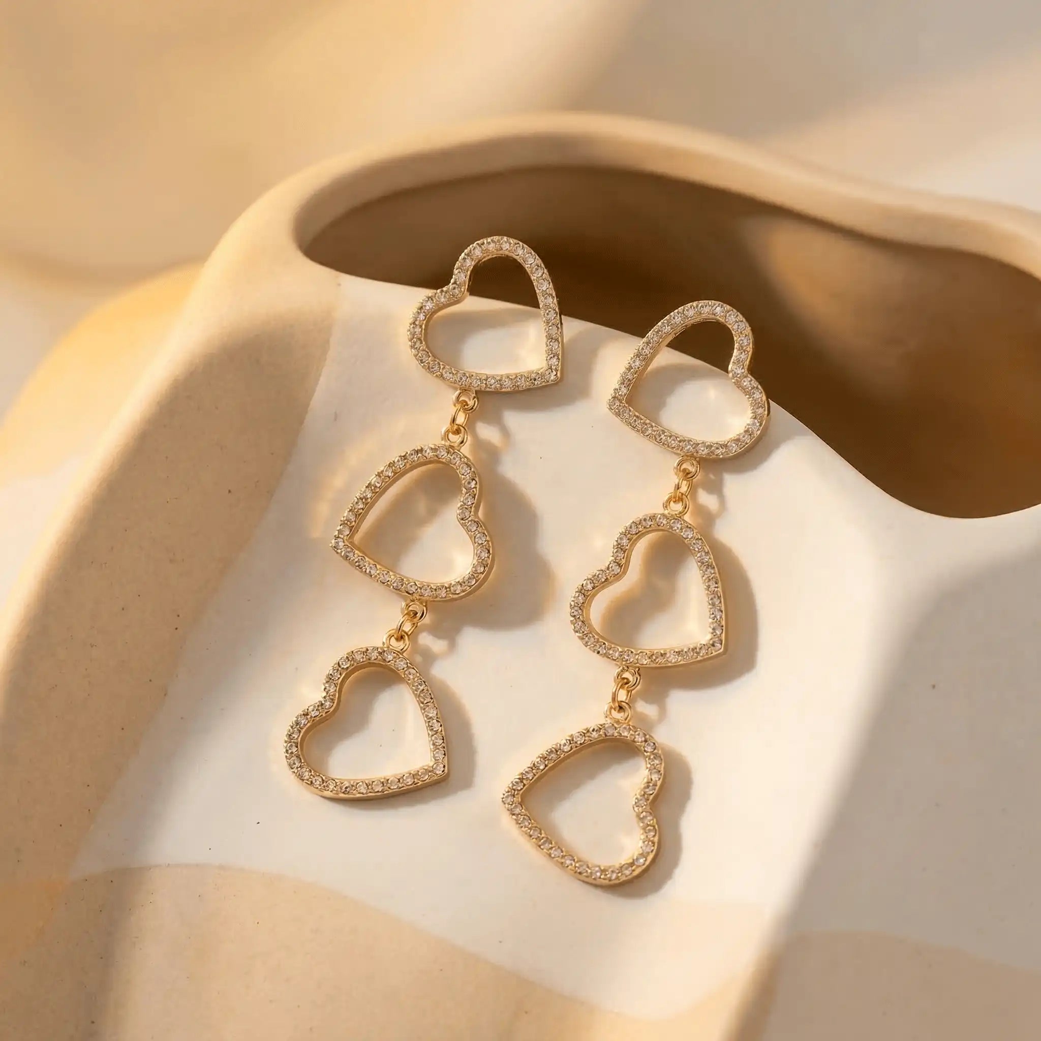 Three-Tier Heart Earrings featuring three linked open-heart shapes, each outlined with small sparkling stones, displayed on a soft beige ceramic surface in warm lighting.