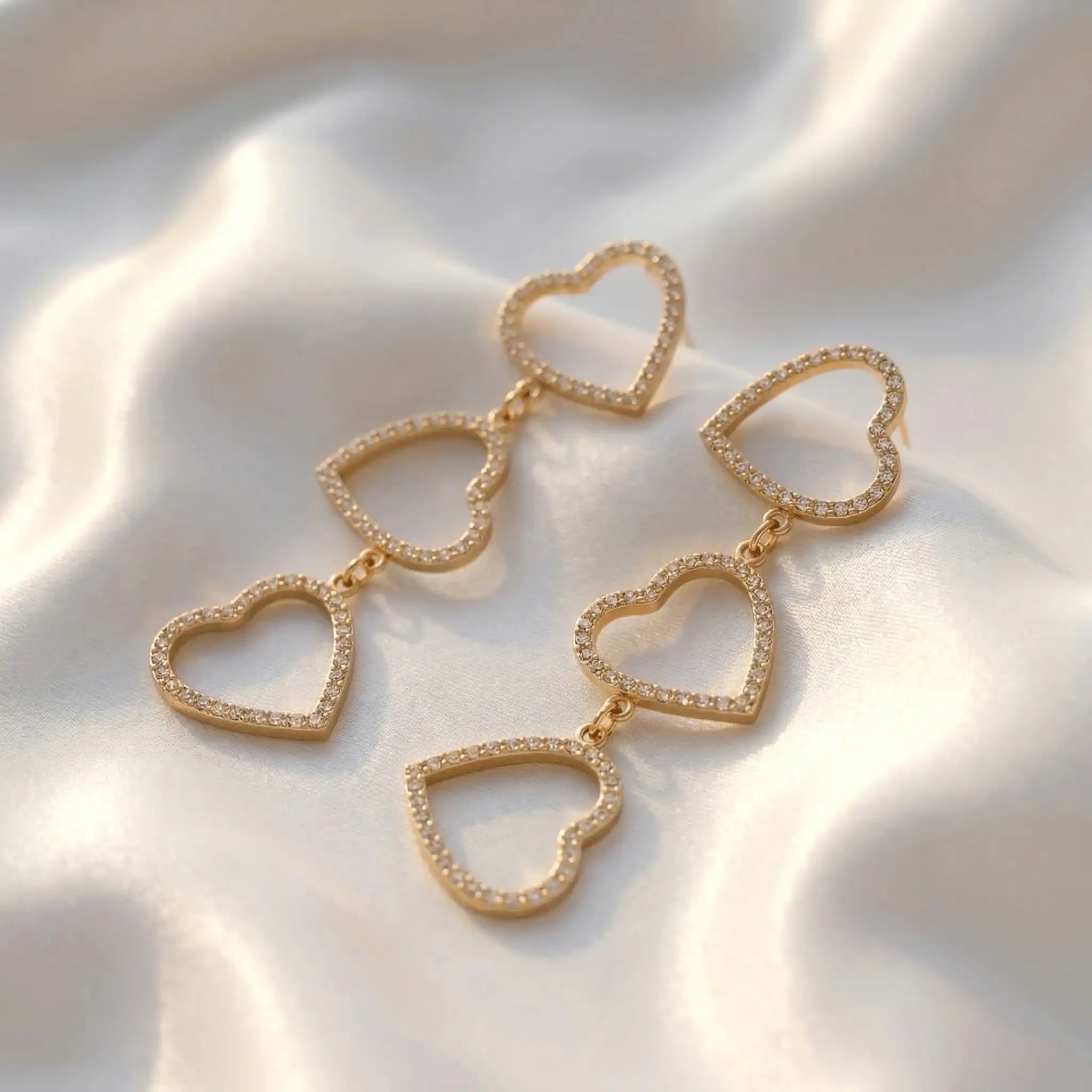 Three-Tier Heart Earrings with cascading open-heart designs and shimmering accents, resting on silky white fabric with gentle folds and highlights.