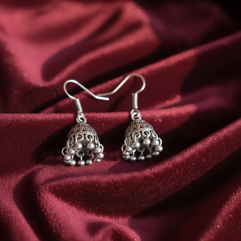 Pair of tiny oxidised jhumkas placed on rich maroon fabric, highlighting their detailed dome design and hanging beads.