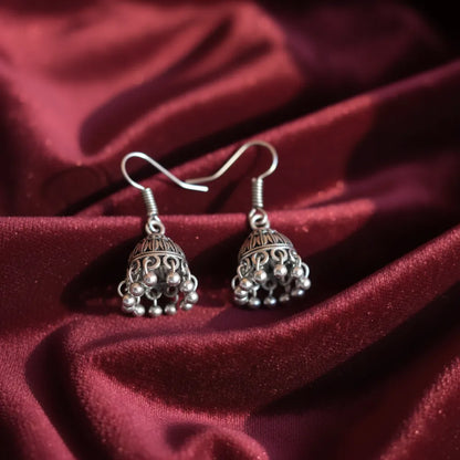 Pair of tiny oxidised jhumkas placed on rich maroon fabric, highlighting their detailed dome design and hanging beads.