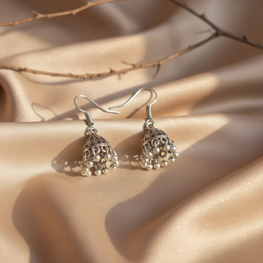 Tiny oxidised jhumkas displayed on soft beige satin fabric with natural light, showing their delicate handcrafted details.