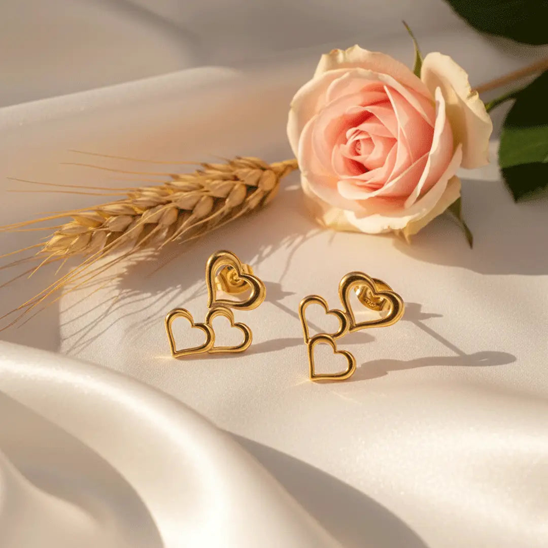 Triple Heart Drops gold earrings placed on white satin with a pink rose and wheat stalk in soft sunlight.