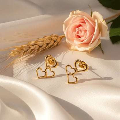 Triple Heart Drops gold earrings placed on white satin with a pink rose and wheat stalk in soft sunlight.