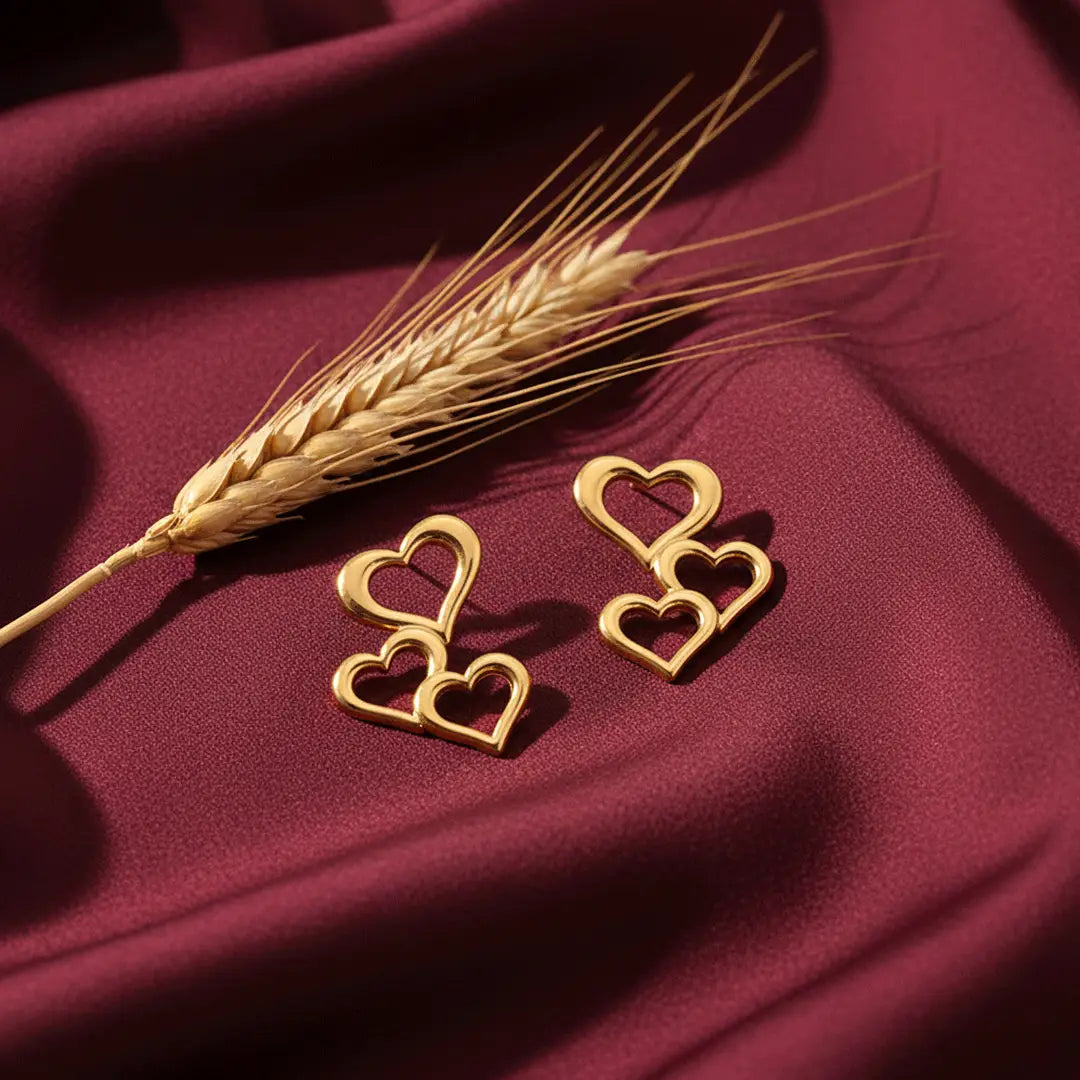 Triple Heart Drops gold earrings displayed on maroon satin fabric with a wheat stalk in soft sunlight.