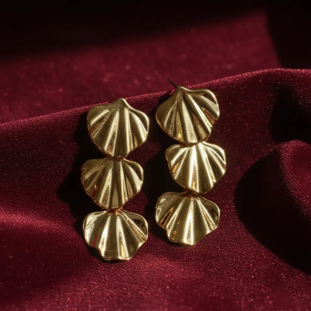 Gold triple-shell statement earrings placed on deep red velvet fabric with soft natural light highlighting their textured design