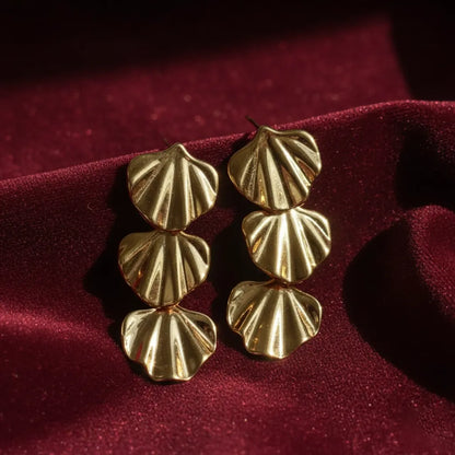 Gold triple-shell statement earrings placed on deep red velvet fabric with soft natural light highlighting their textured design