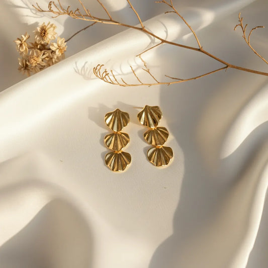 Gold triple-shell statement earrings displayed on white satin fabric with dried flowers and warm sunlight creating an elegant aesthetic.