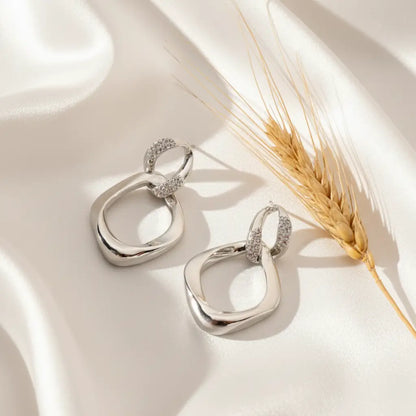Silver abstract drop earrings with twisted loop design placed on soft white satin fabric with a wheat stem beside them.