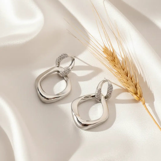 Silver abstract drop earrings with twisted loop design placed on soft white satin fabric with a wheat stem beside them.