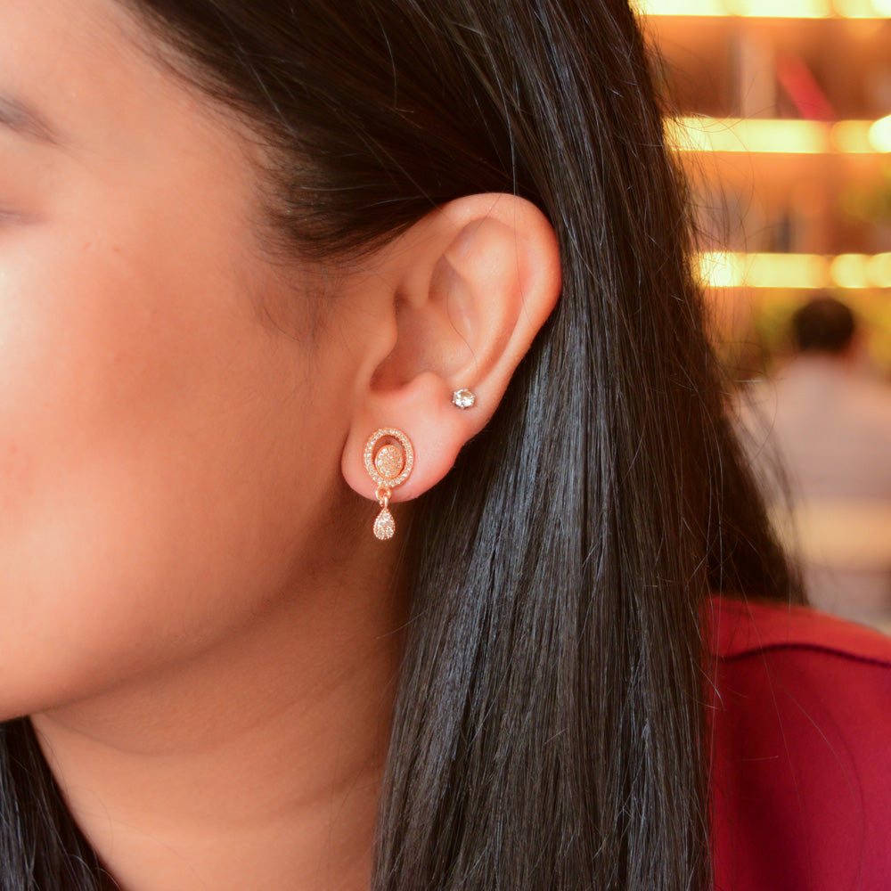 A model wearing Twinkle Drop AD Earrings in rose gold, featuring a circular crystal-studded design with a dangling teardrop stone.