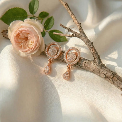 Twinkle Drop AD Earrings in rose gold with circular crystal tops and teardrop dangles, styled on ivory satin beside a soft pink rose and a decorative twig.