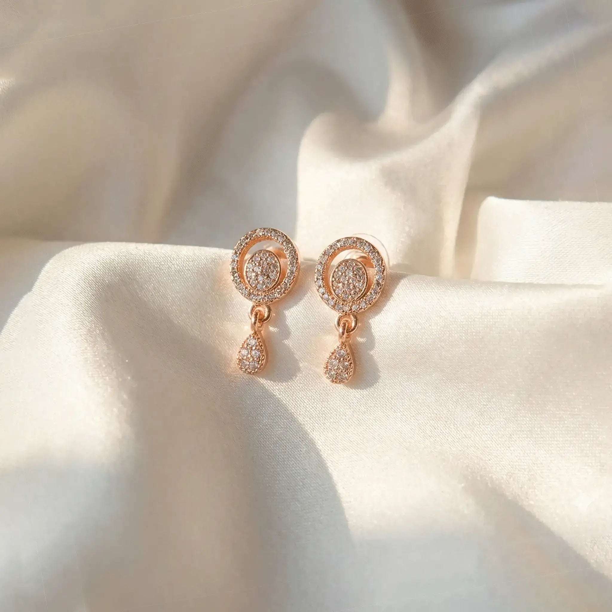Twinkle Drop AD Earrings in rose gold, featuring a circular crystal-studded design with a dangling teardrop stone, displayed on soft ivory satin fabric.