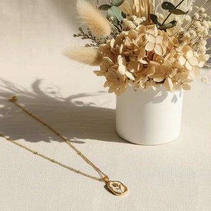 Gold White Rose Pendant necklace placed on a soft beige surface beside a vase of dried flowers.