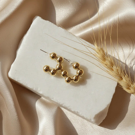Golden beaded earrings arranged in a loop shape, displayed on a white textured stone over beige satin fabric with wheat decor.