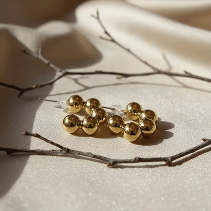 Beaded golden earrings with polished round beads placed on soft beige fabric with natural twig accents.